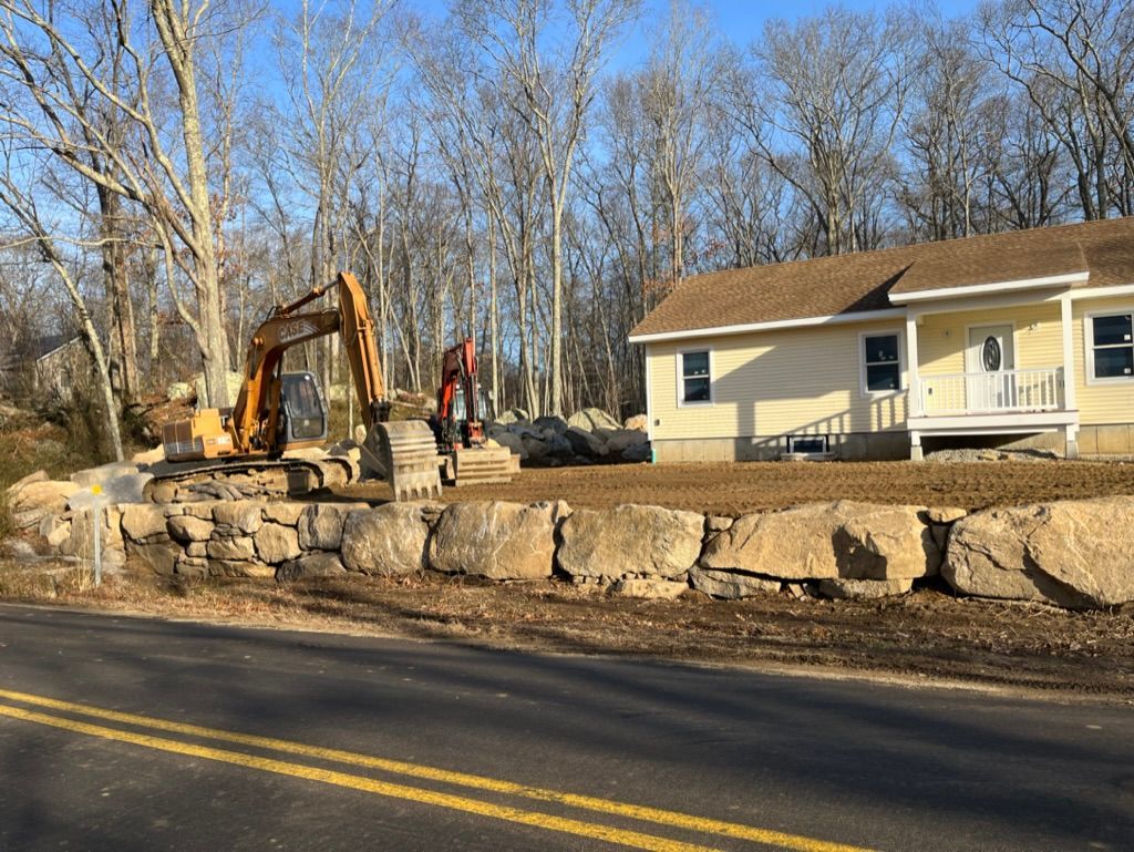 Excavator building a stone retaining wall in front of a yellow house with a brown roof.