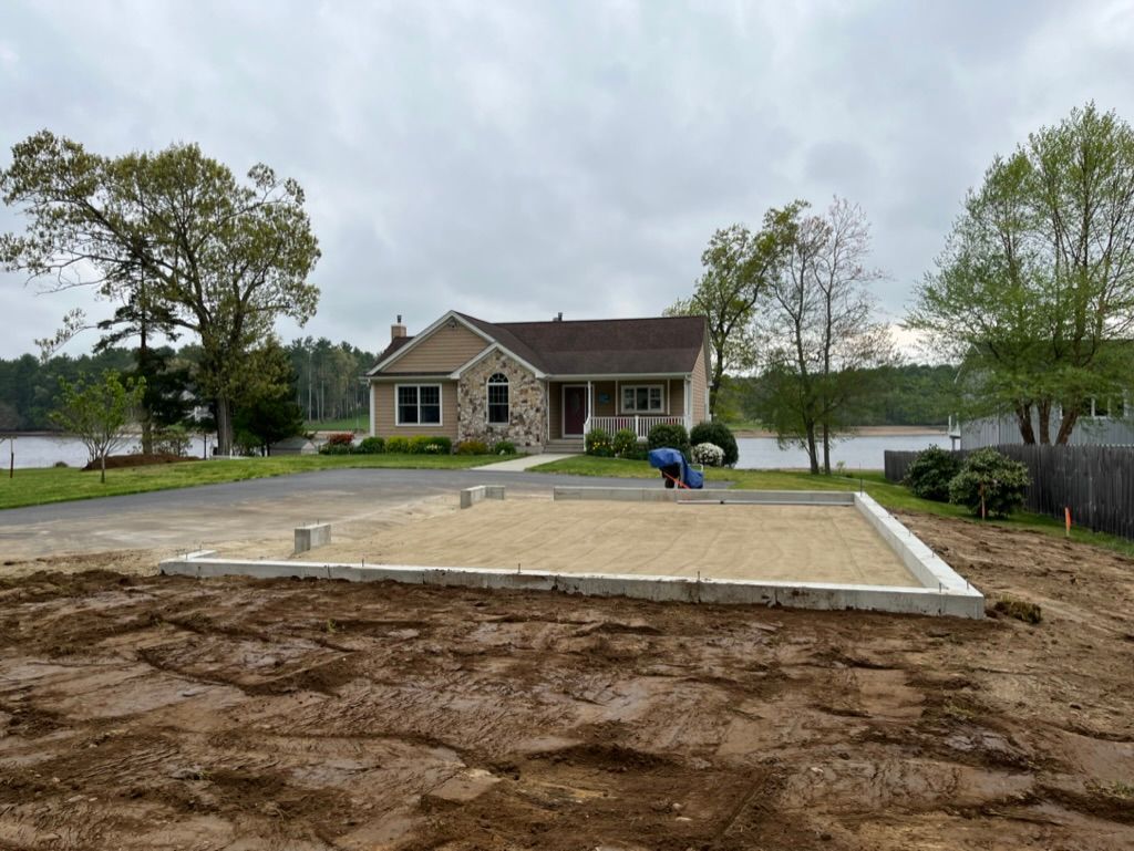 House under construction. Concrete foundation in foreground, house with stone facade in background, cloudy sky.