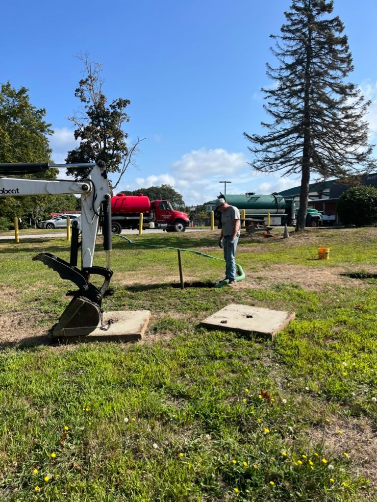 Man vacuuming a septic tank. Two concrete lids lie open. A truck is in the background. Sunny day.