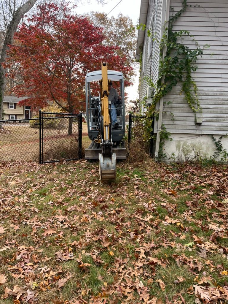Mini excavator operating near a house, preparing to dig. Fall foliage and a black fence are visible.