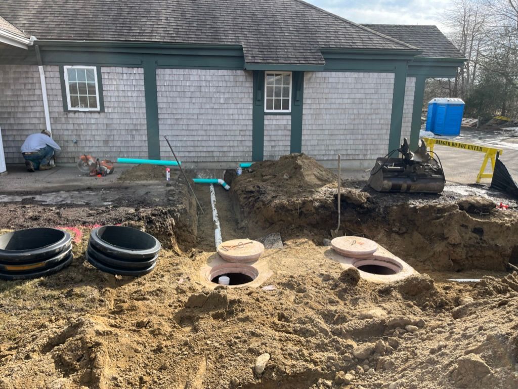 Construction site: septic tank installation. Ground open, pipes, tanks visible; worker, building exterior.