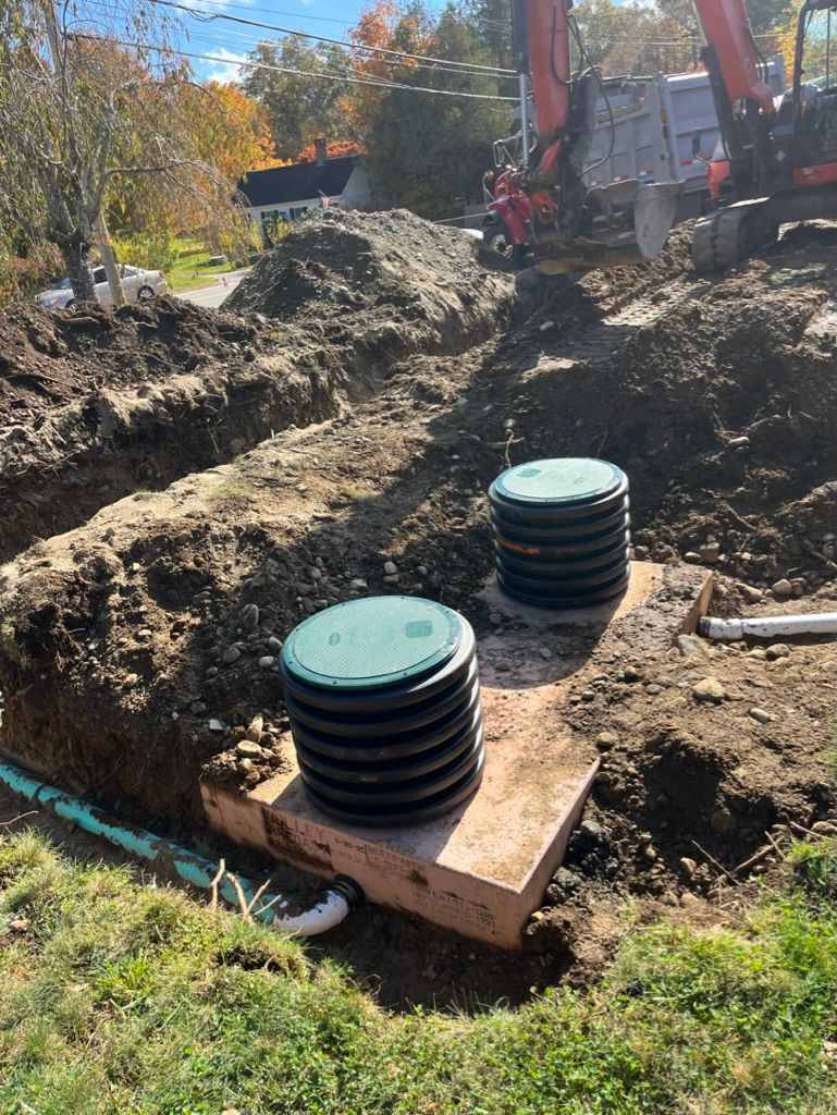 Construction site with excavated trench, two septic tank covers, and an excavator in the background.