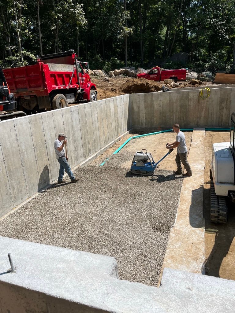 Construction workers compacting gravel base in a concrete foundation pit with heavy machinery; red dump truck in background.