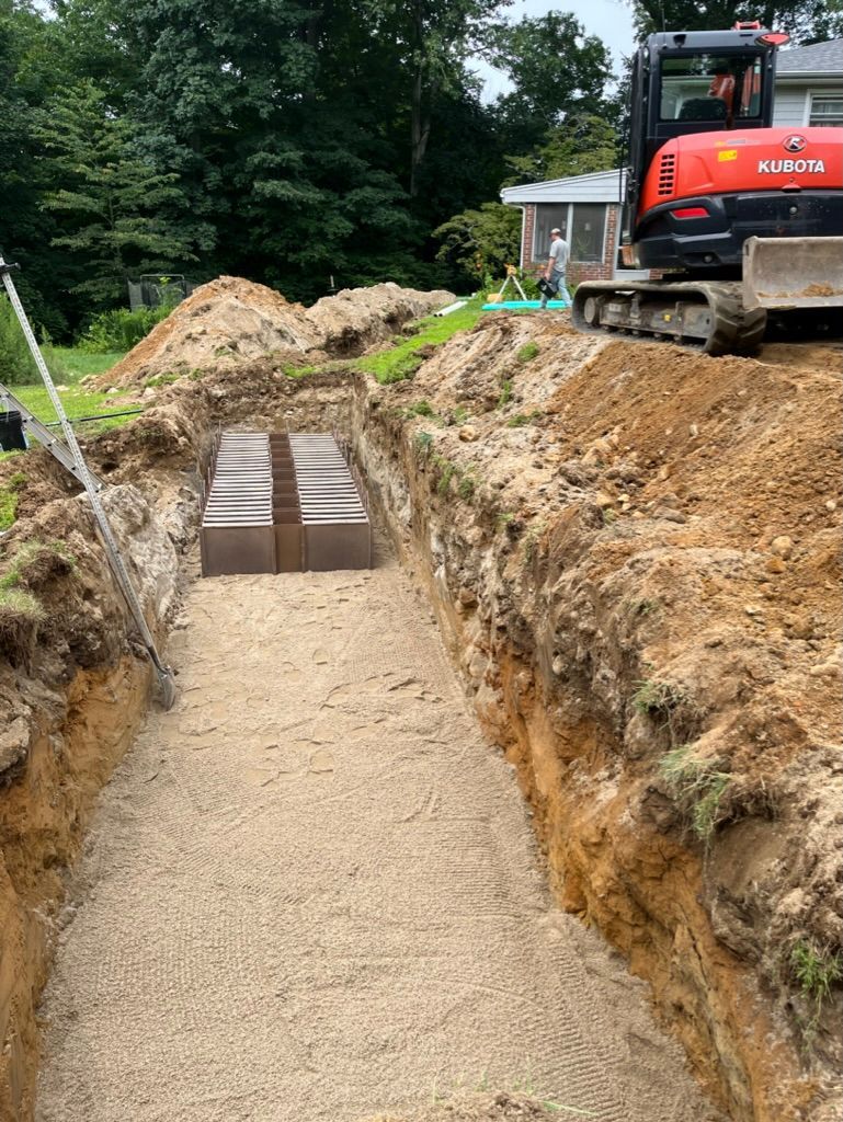 Excavation site with a long, sandy trench. A backhoe sits nearby. A structure is partially buried.