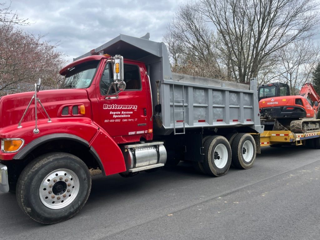 Red dump truck with a flatbed trailer carrying an excavator on a residential street. Cloudy sky.