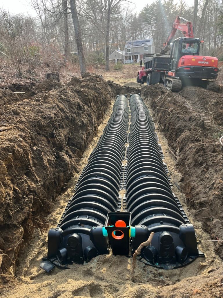 Trench with plastic septic chambers. An excavator and dump truck are in the background, near a wooded area.