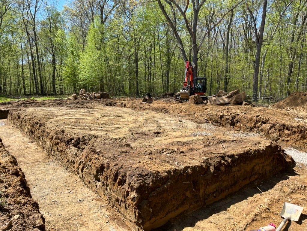 Excavated rectangular foundation pit; a backhoe and forest backdrop.