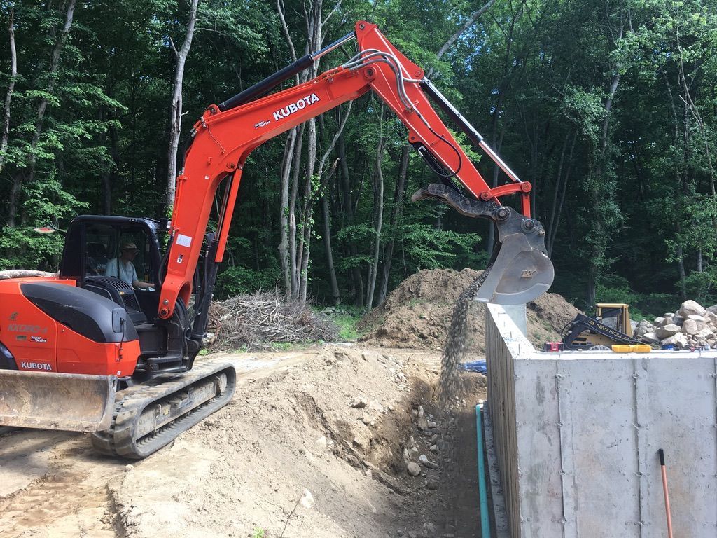 An orange Kubota excavator digs soil next to a concrete wall in a wooded area.