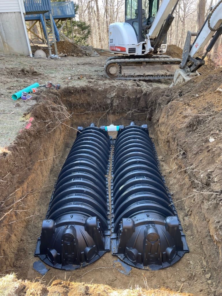 Two connected black plastic septic tanks installed in a dirt excavation, with a backhoe in the background.