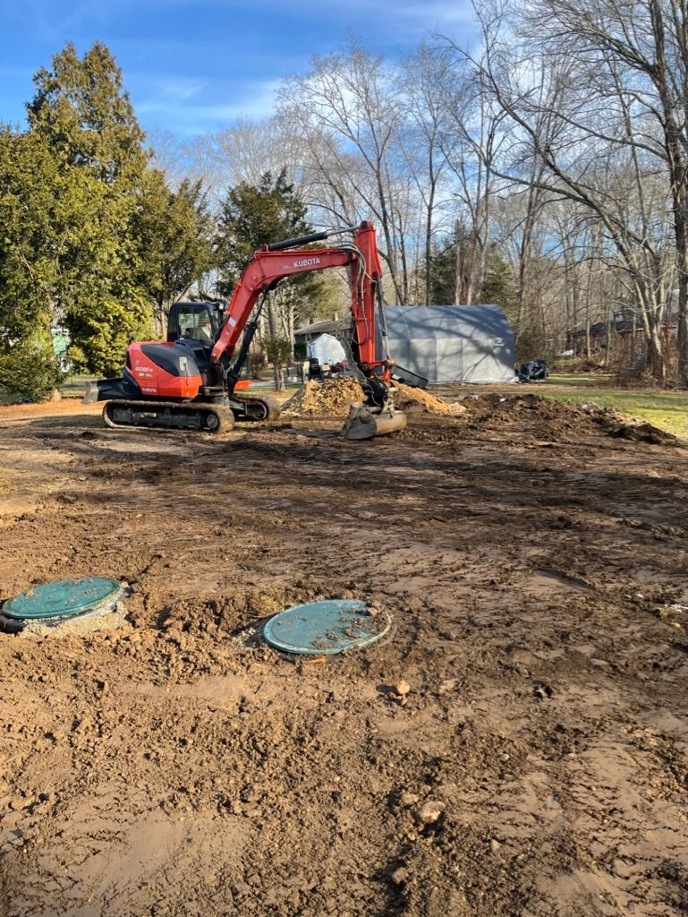Excavator digging in muddy ground near two septic tank lids. Trees and a building in the background.