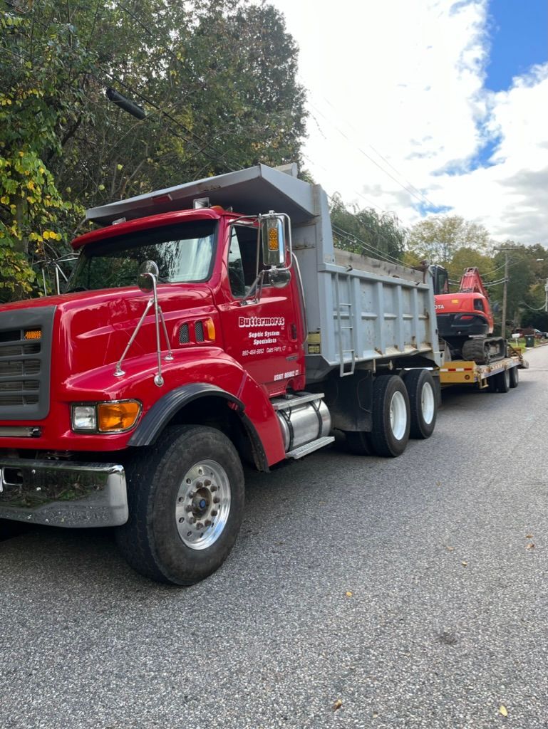 Red dump truck with a trailer carrying an excavator on a paved road.
