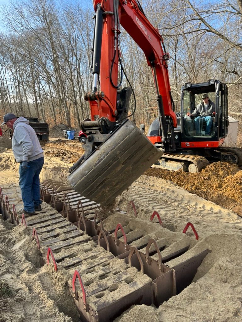 An excavator pours dirt into a trench lined with metal forms. A man observes. Outdoors.