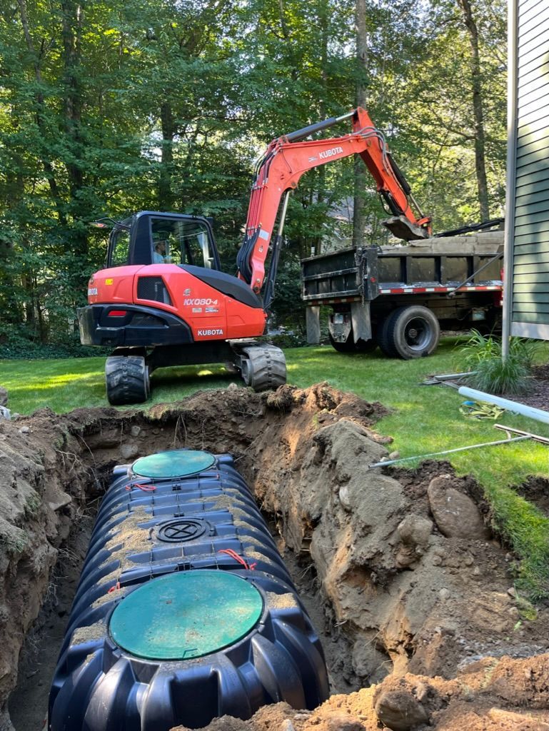An excavator and dump truck are placing soil on a septic tank in a yard.