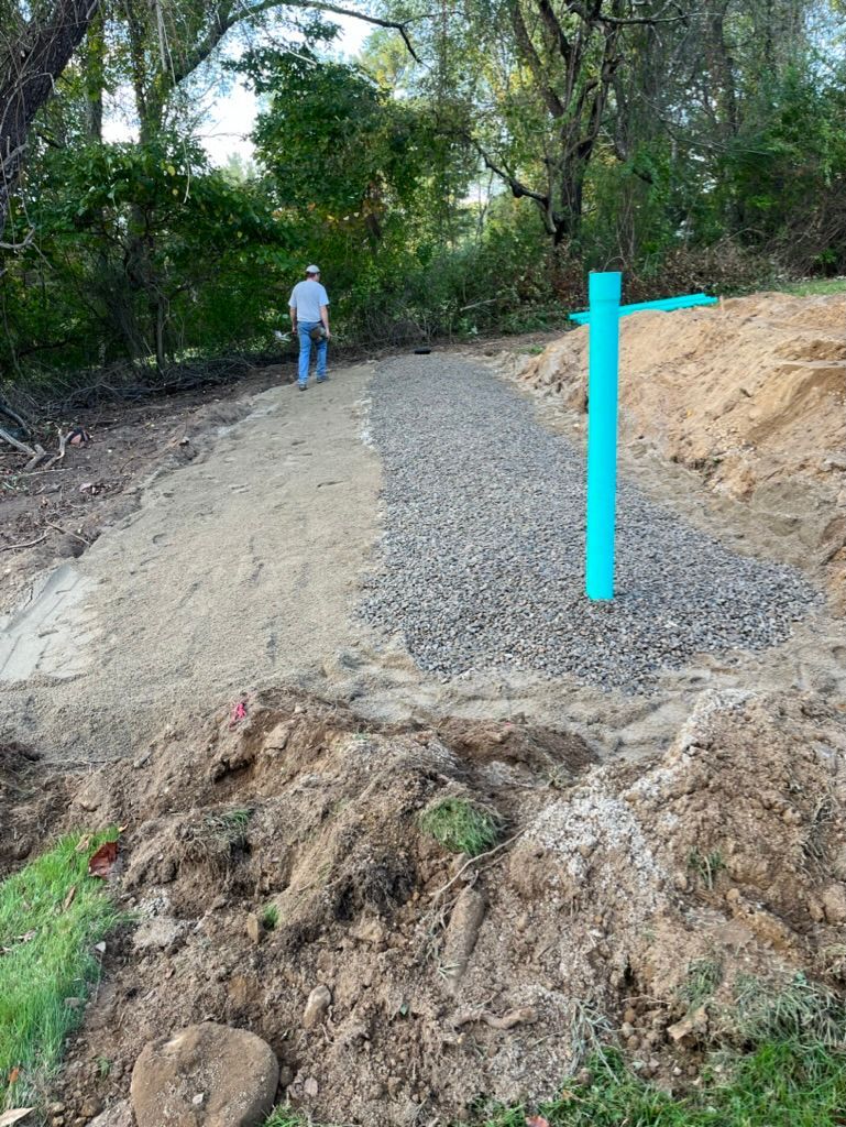 A gravel path through a wooded area; a person walks away. Turquoise pipe stands at edge.