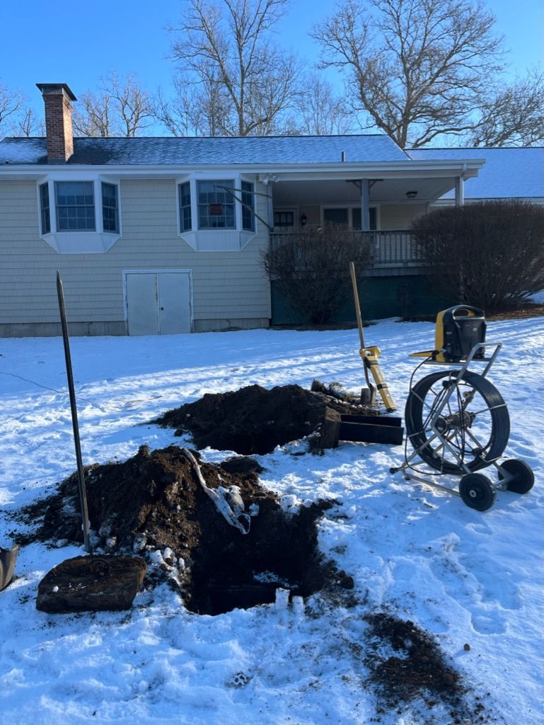 Hole dug in snow in front of a house. Black dirt pile and equipment for inspecting pipes.