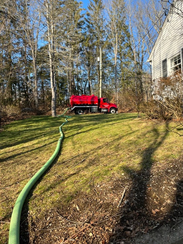 Red septic tank truck with green hose on a lawn; tall trees in background next to a house.
