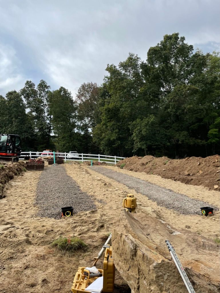 Construction site with gravel pathways, dirt, and equipment; white fence and trees in the background.