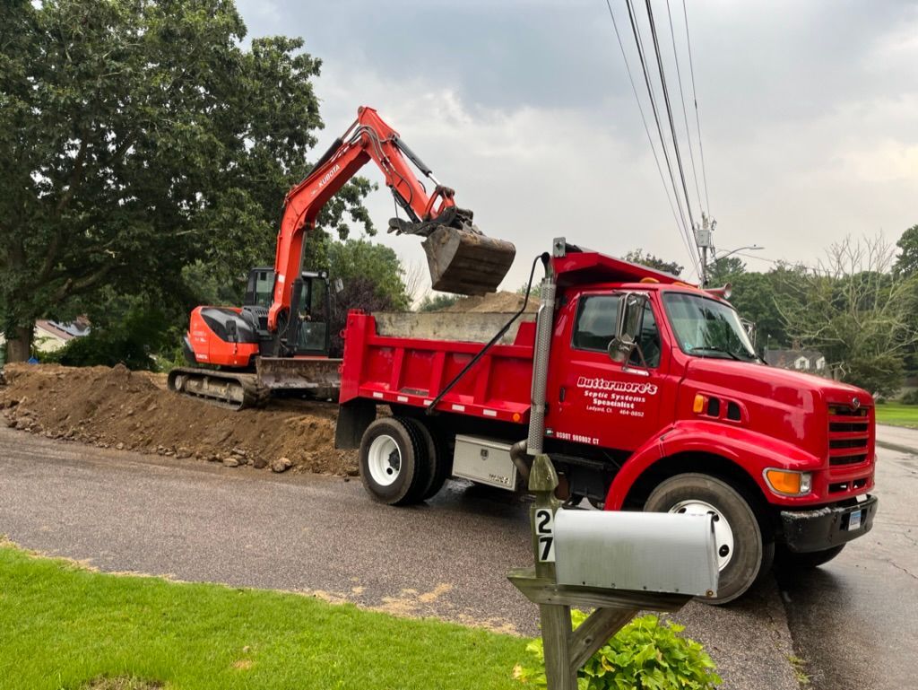 An excavator loading dirt into a red dump truck on a street.
