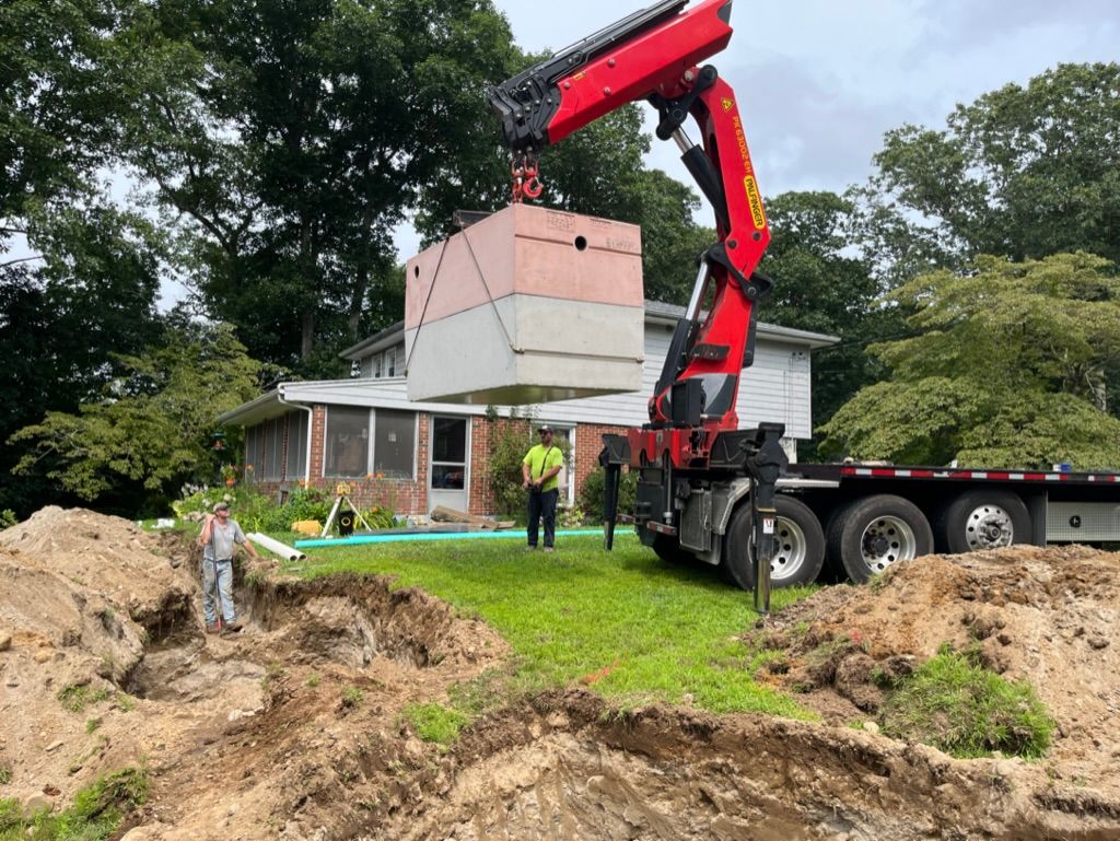 A crane lifts a large concrete structure over a house. Two workers supervise the operation in a yard.