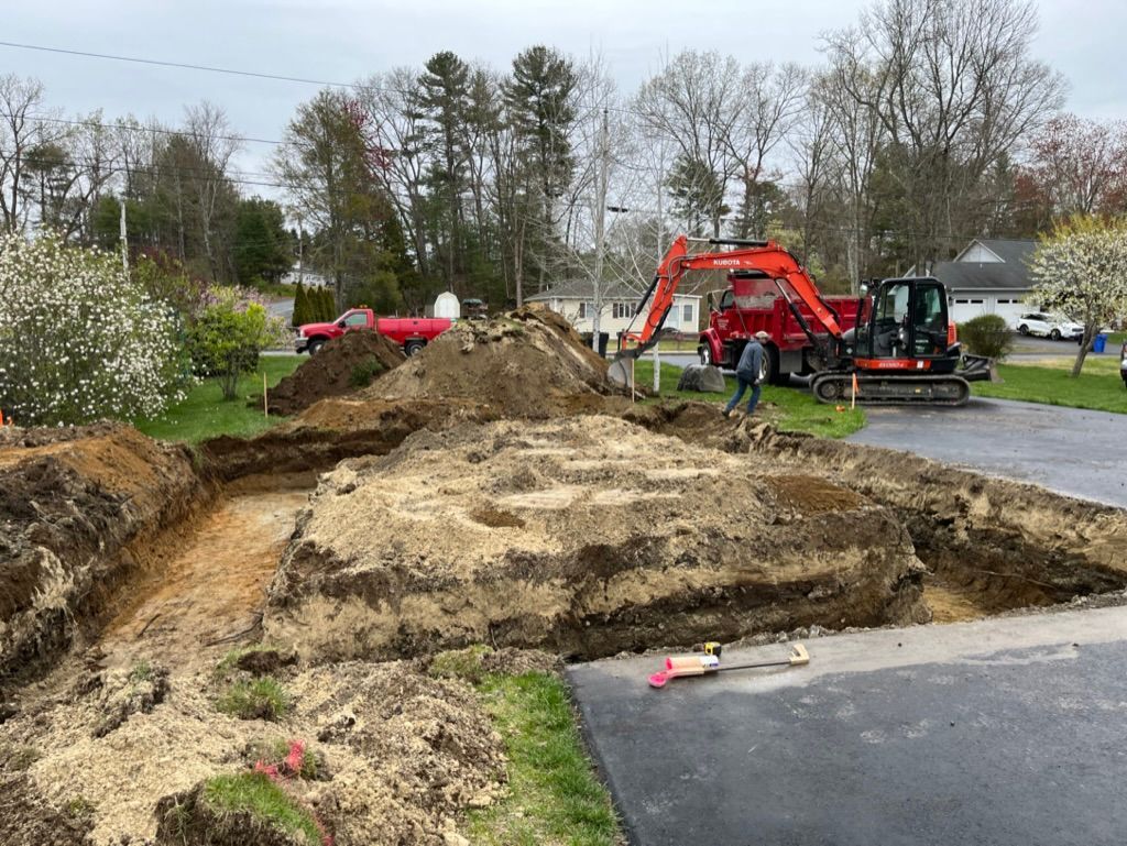 Construction site with excavator digging a foundation in a residential area. Dirt piles, asphalt driveway.