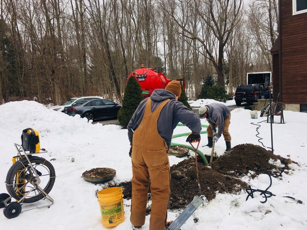 Two people digging in snowy yard; one wears brown overalls, green hose visible, brick house in background.