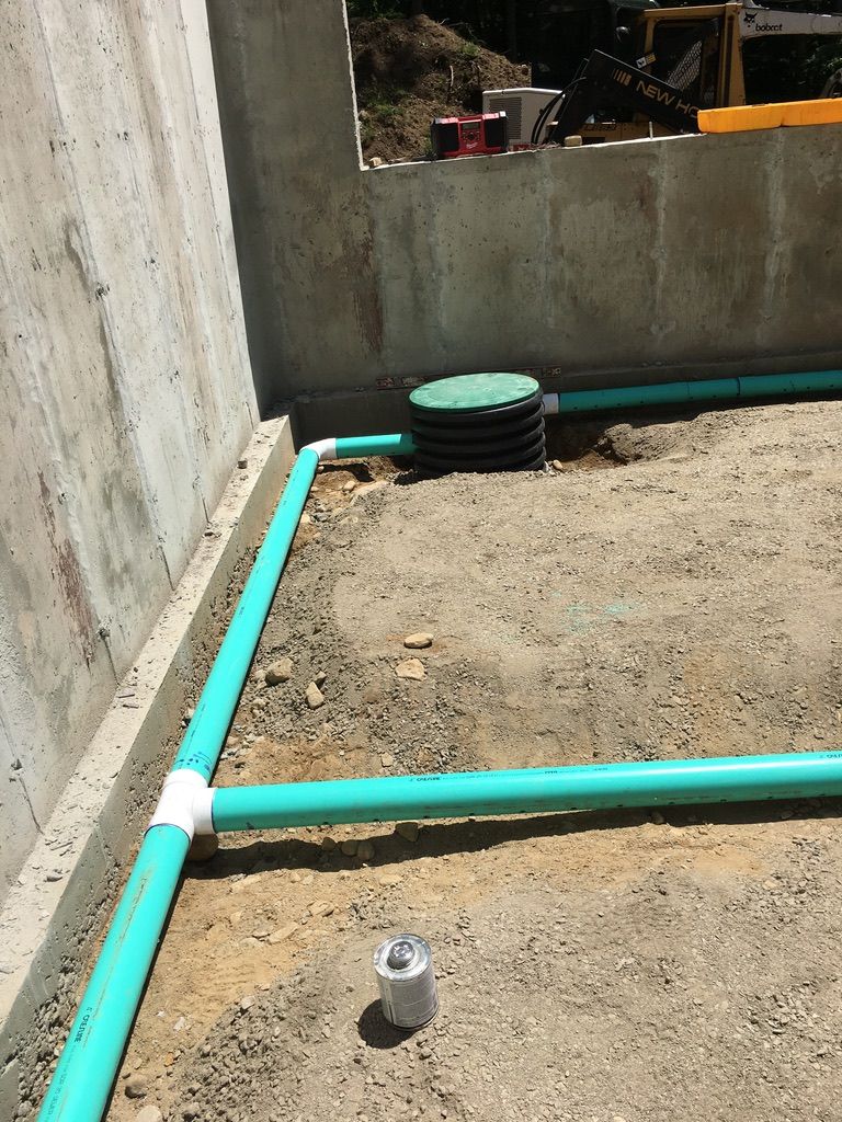 Green plumbing pipes installed in a dirt-floored construction site near a concrete wall.