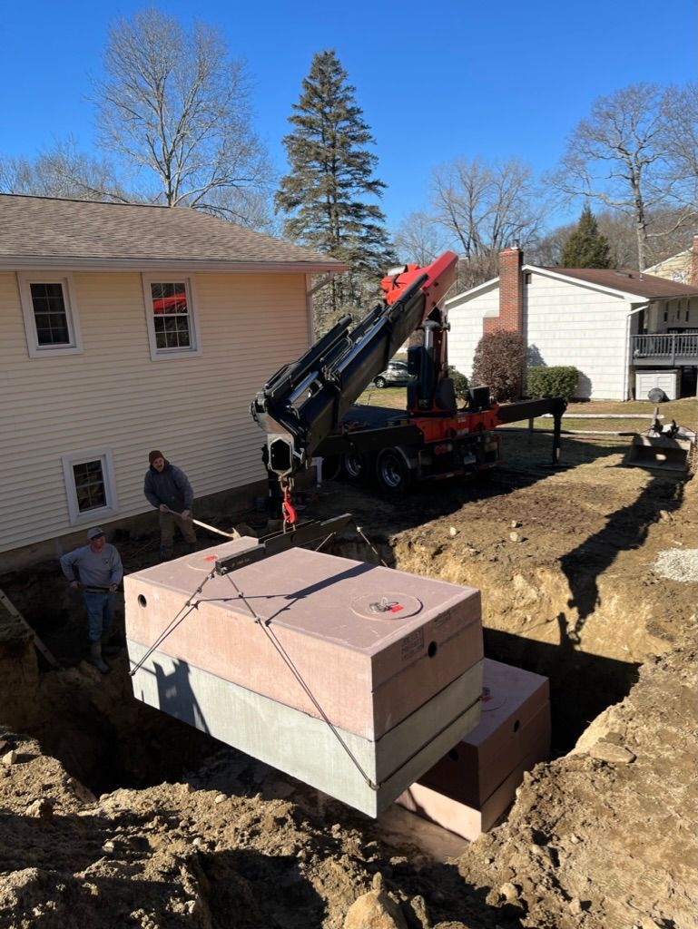 A crane lowers a concrete septic tank into an excavated trench beside a house.