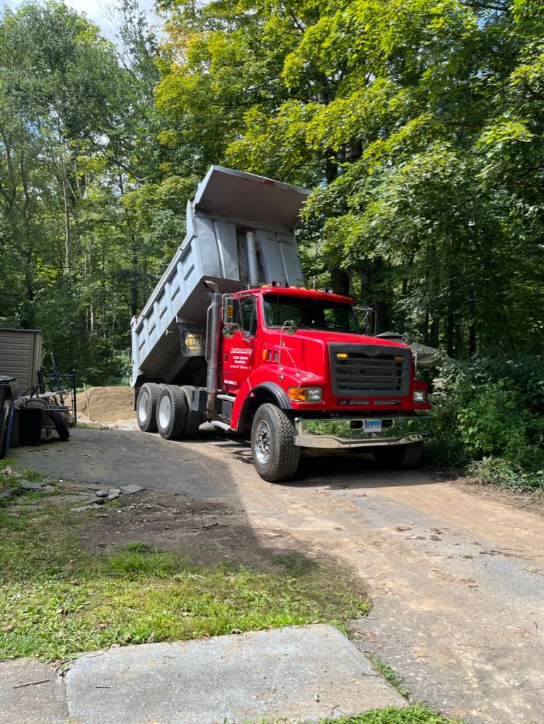 Red dump truck with raised bed on a dirt driveway, unloading material. Trees in background.