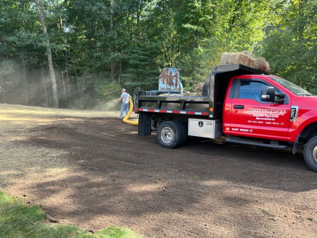 Red truck spreading mulch in a yard. A person in a mask operates a machine, and trees are in the background.