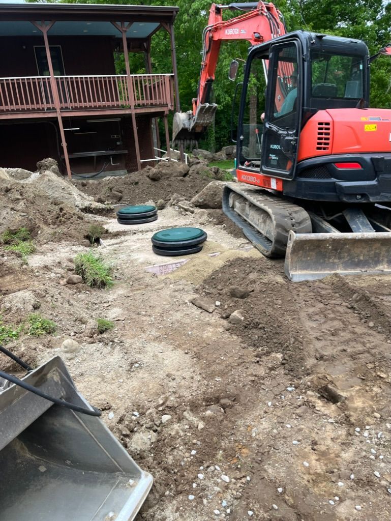 Excavator near a house with a deck. Two green septic tank covers are in the dirt.