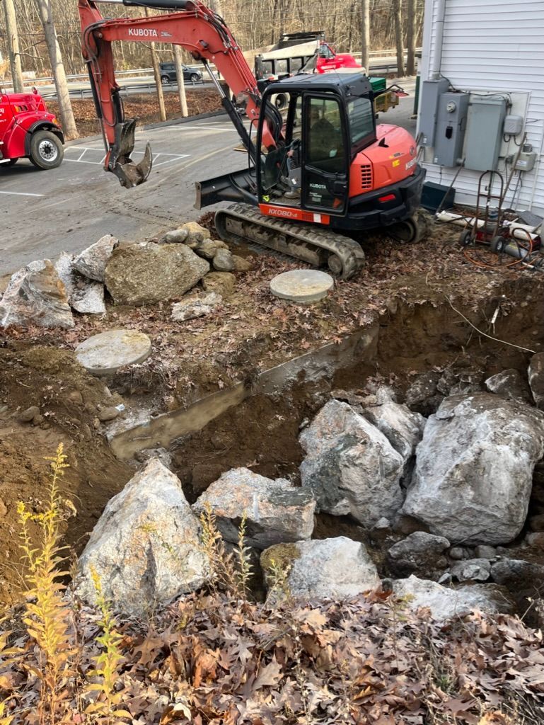 An excavator removing rocks and soil near a building. Outdoor setting, overcast.