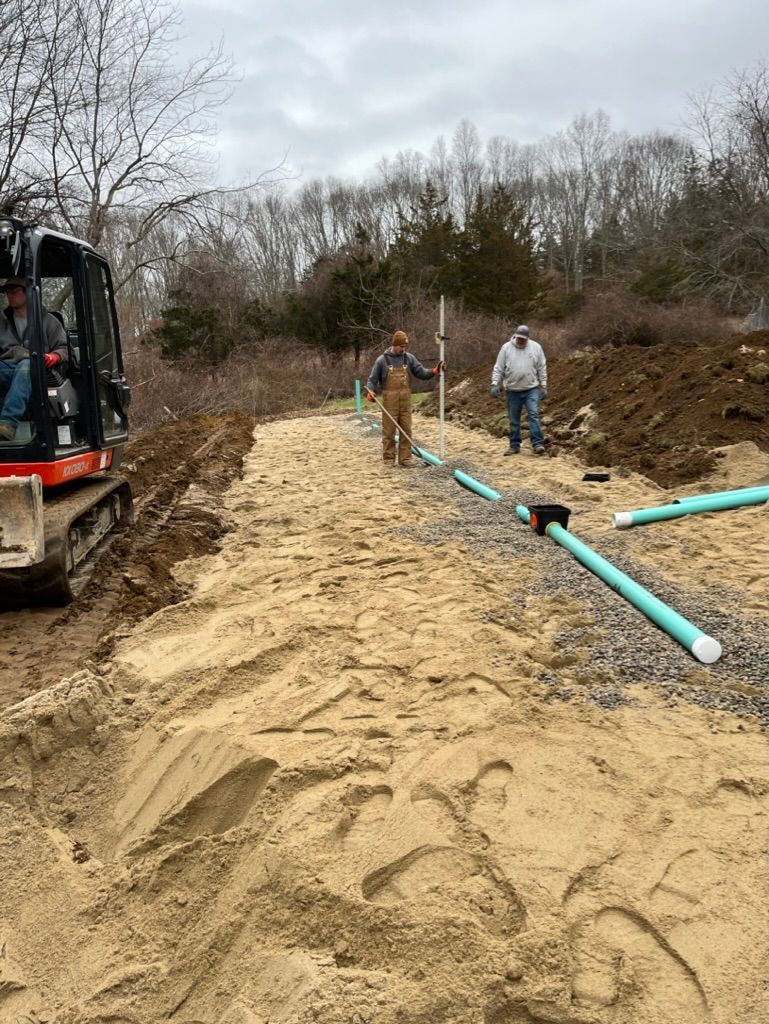 Construction site: men installing pipes on sand. Excavator on the left, trees in background. Cloudy day.