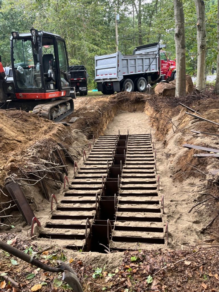 Construction site: excavator and dump truck over an open trench with metal grates installed.