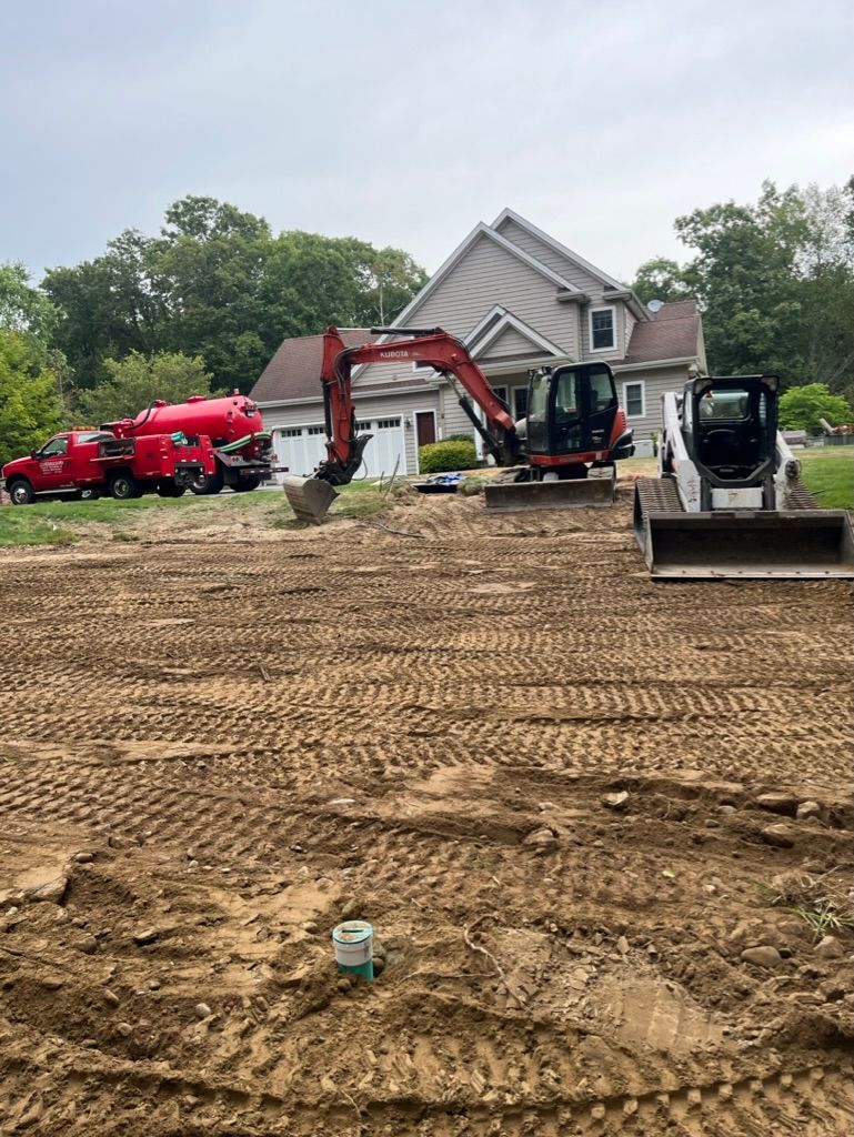 Construction site with excavator and skid steer in front of a house; red trucks parked nearby.