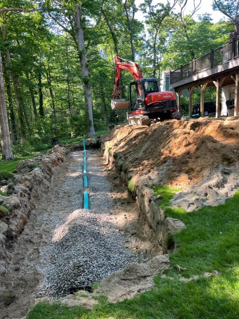 Excavator digging a trench for pipe installation near a house in a wooded area; gravel and dirt visible.
