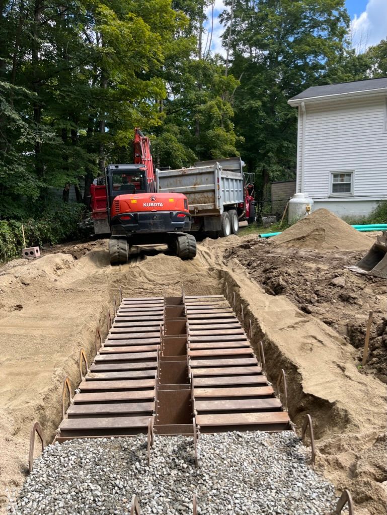 Construction site with an excavator, dump truck, and trench lined with wood and gravel.
