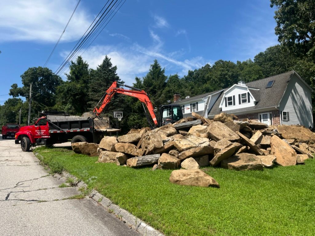 A red excavator and dump truck removing a pile of rocks from a grassy area next to a building on a sunny day.