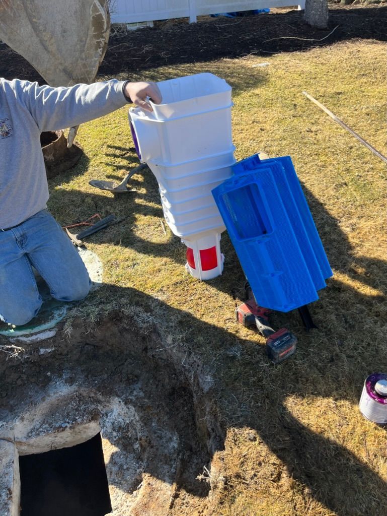 Person holding a white chicken feeder and blue waterer on grass. A drill sits nearby.