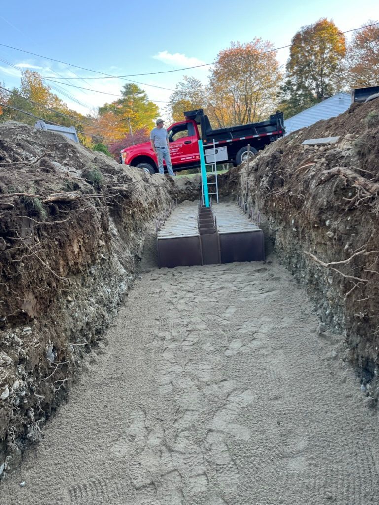 Dug trench with gravel base; metal rectangular structure centered; red truck and worker in the background.