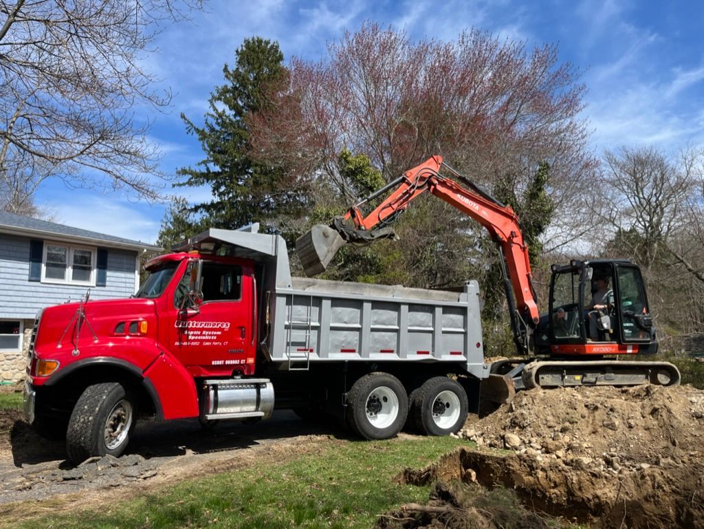A red dump truck being loaded with dirt by a small excavator in a residential yard.