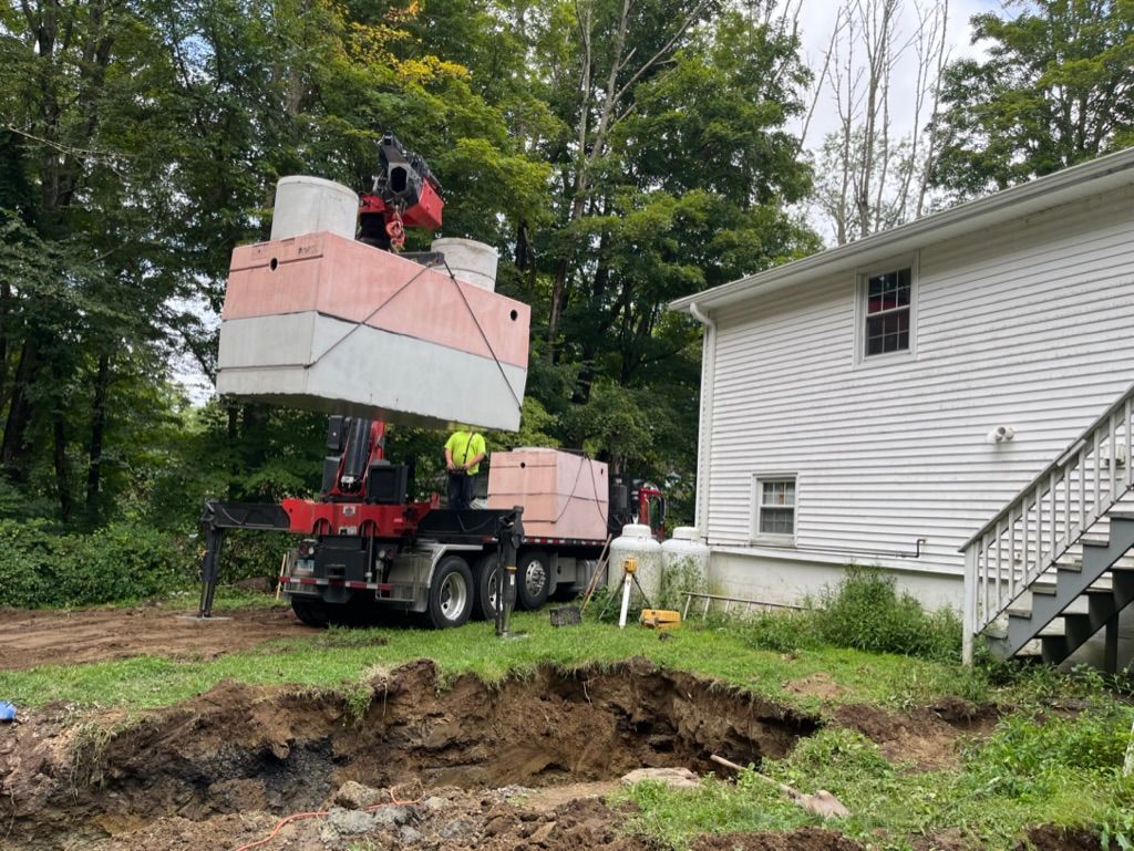 A crane lifting a large septic tank over a dug hole next to a white house.