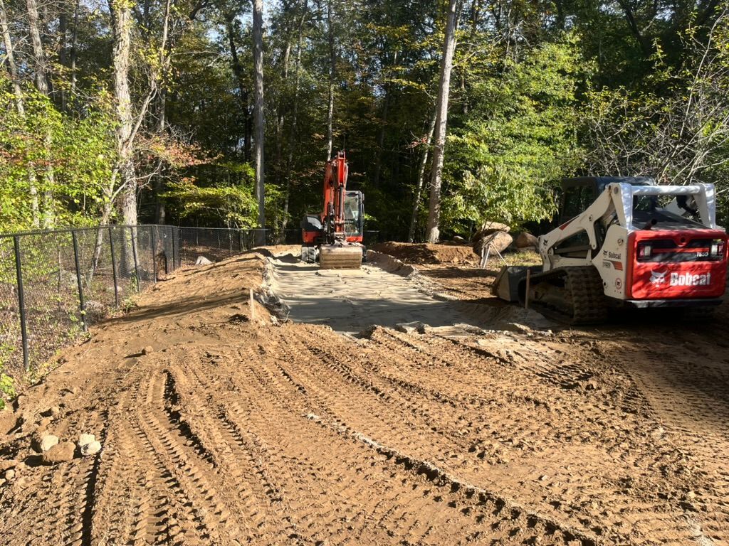 Construction site with excavator and bobcat. Dirt piles, trees, and a fence are also visible.