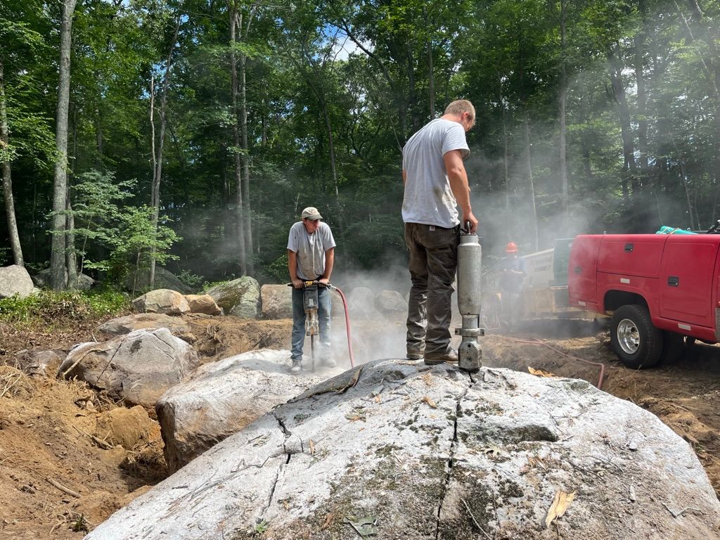 Two workers use jackhammers to break a large rock. Dust is in the air; red truck is visible.