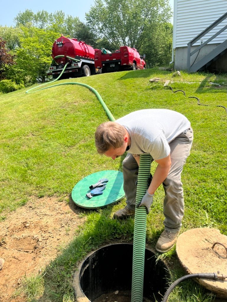 Man emptying septic tank with a hose. Red tank truck in the background, green grass, and a sunny day.