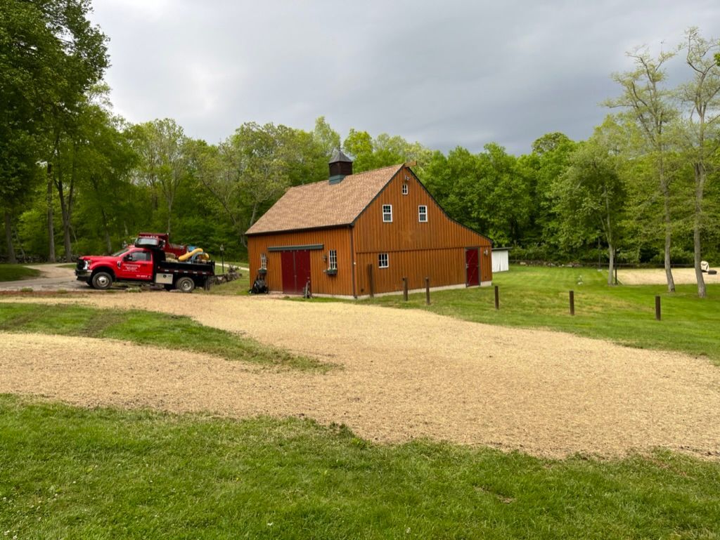 Red pickup truck parked near a brown barn with a dark roof on a gravel drive in a wooded area.