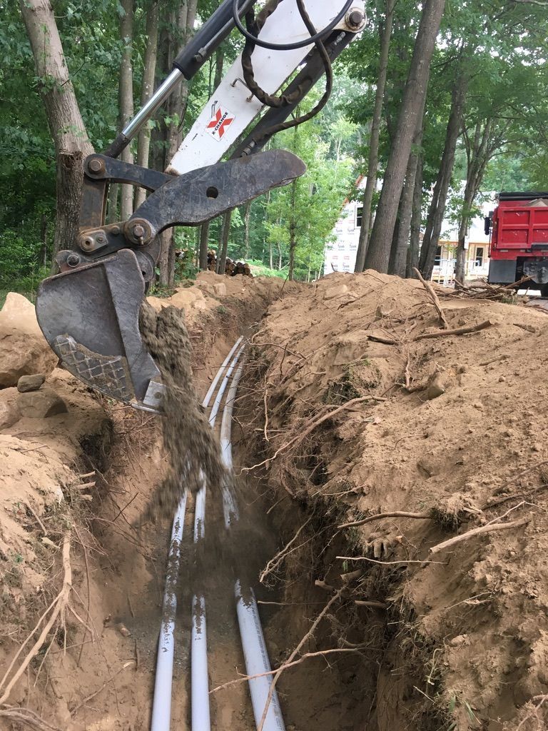 Mini-excavator backfills a trench containing several white pipes, near trees and a red truck.