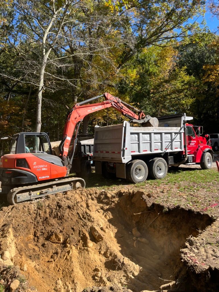 Orange excavator loading a red dump truck with dirt in a wooded area.