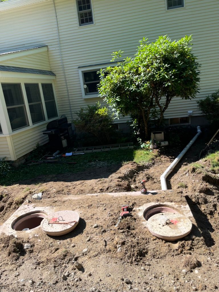 Septic tank access points in a yard. Open lids, exposed ground, house and trees in background.