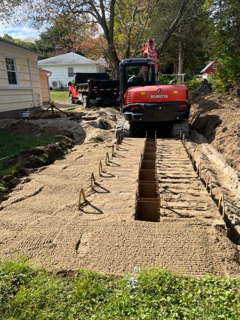 Excavator and trailer on a construction site; trench in sandy soil, residential background.