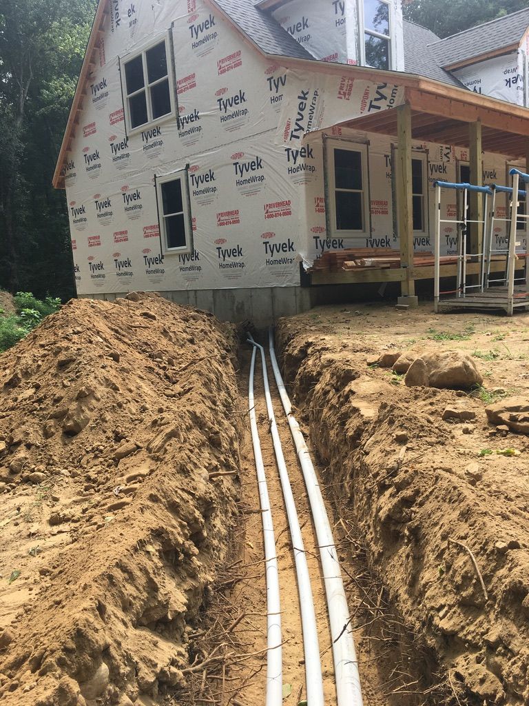 Trench with white pipes leading to a house under construction. Brown soil surrounds them.